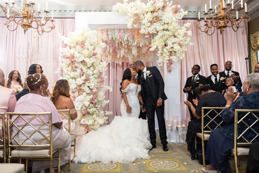 black couple in front of white chuppah covered in blush and ivory flowers sharing their first kiss as man and wife